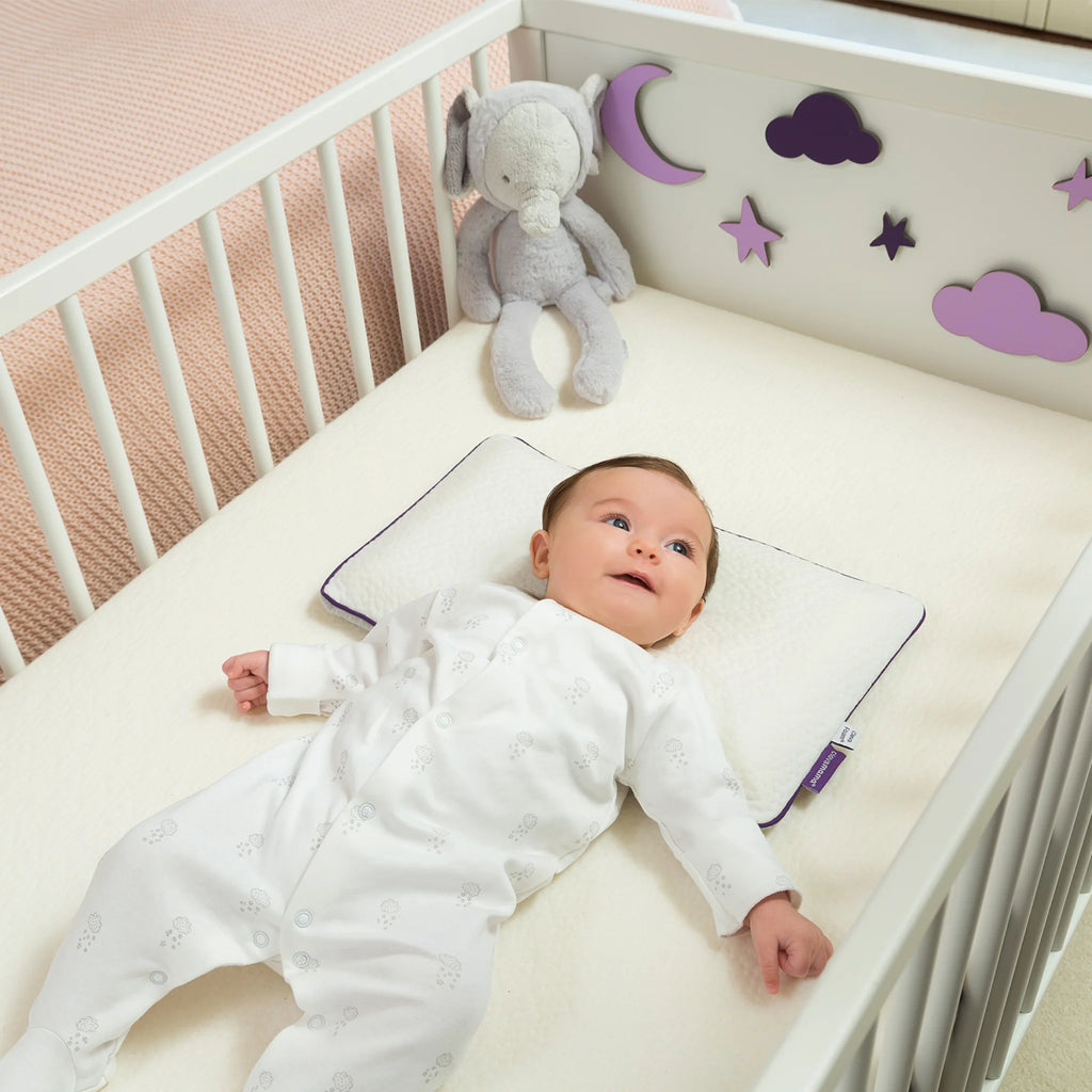 Baby lying in crib on Clevamama ClevaFoam Baby Pillow, ergonomic breathable memory foam pillow with hypoallergenic washable cover, designed for infants 0-12 months to prevent flat head syndrome and support healthy sleep.