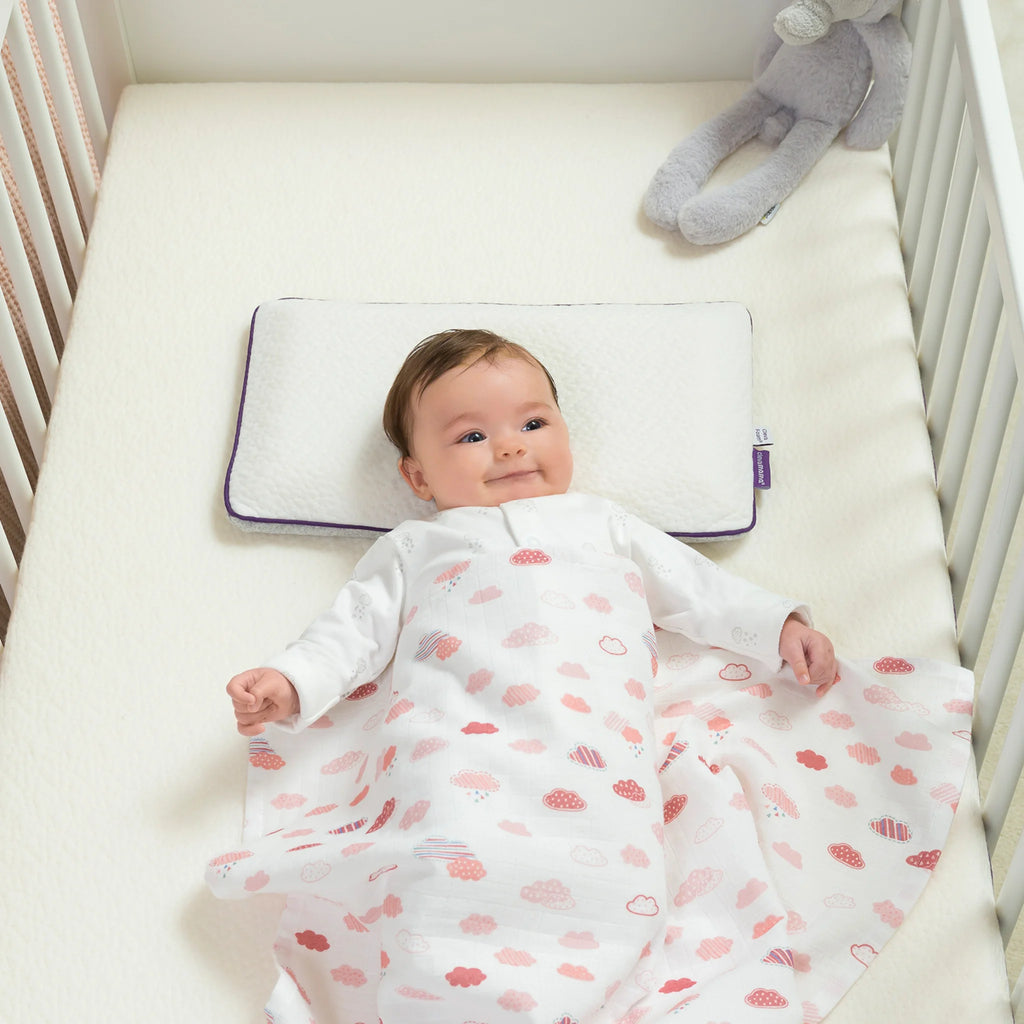 Smiling baby lying in crib on Clevamama ClevaFoam Toddler Pillow, ergonomic breathable memory foam pillow with hypoallergenic washable cover, providing head and neck support for infants and toddlers.