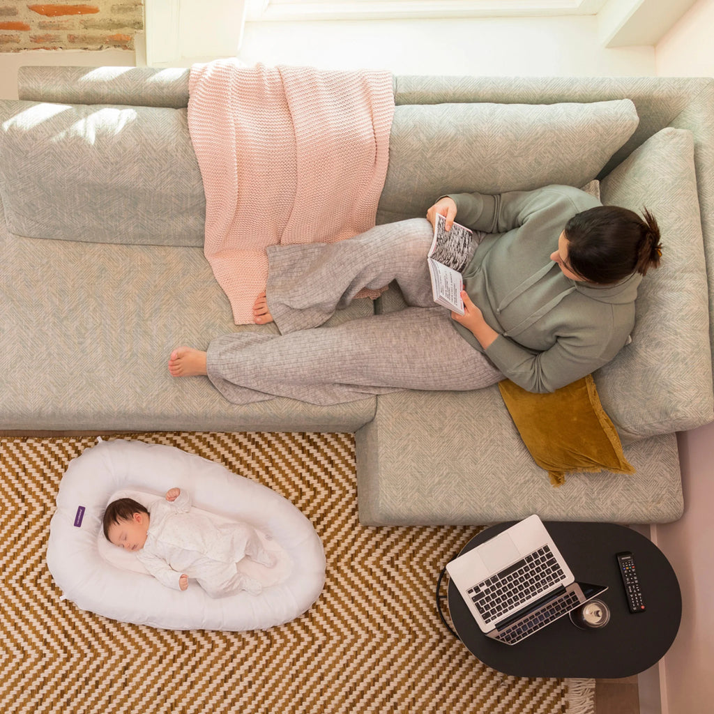 Newborn sleeping in ClevaFoam Baby Pod on living room floor while mother relaxes on sofa ergonomic portable baby lounger for safe supervised rest, comfort and anti-flat head support.”