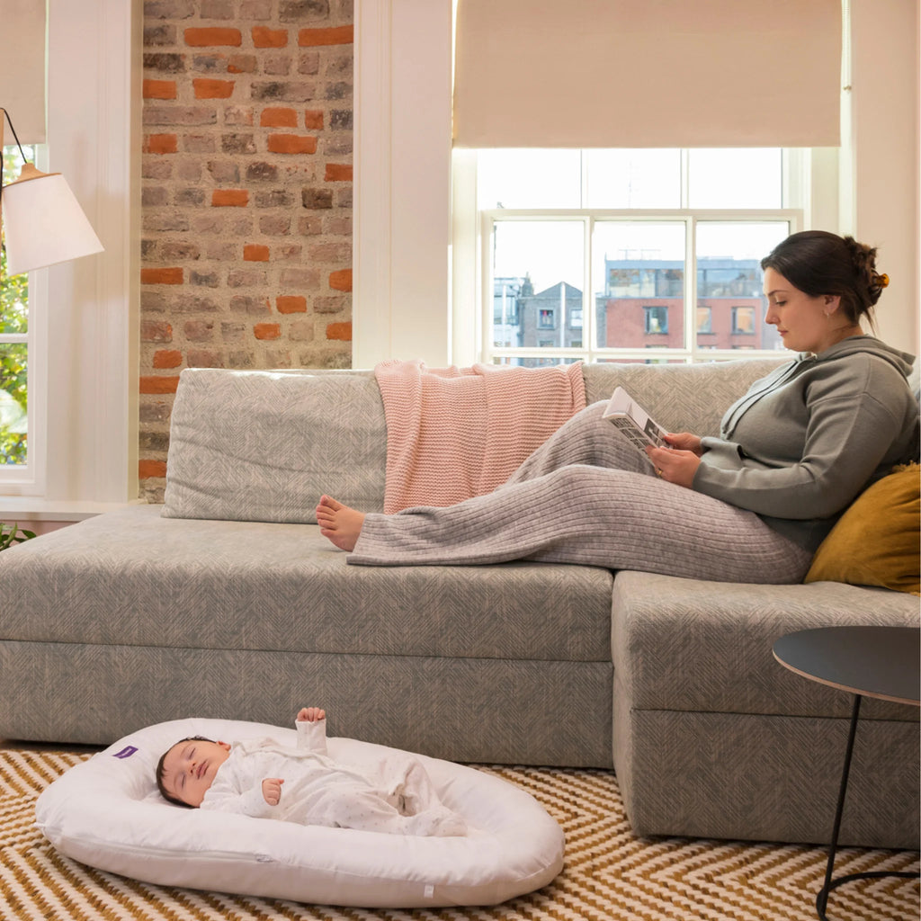 Mother reading on sofa while newborn sleeps in ClevaFoam Baby Pod, ergonomic portable baby lounger with soft padded sides for safe supervised rest, comfort and anti-flat head support.