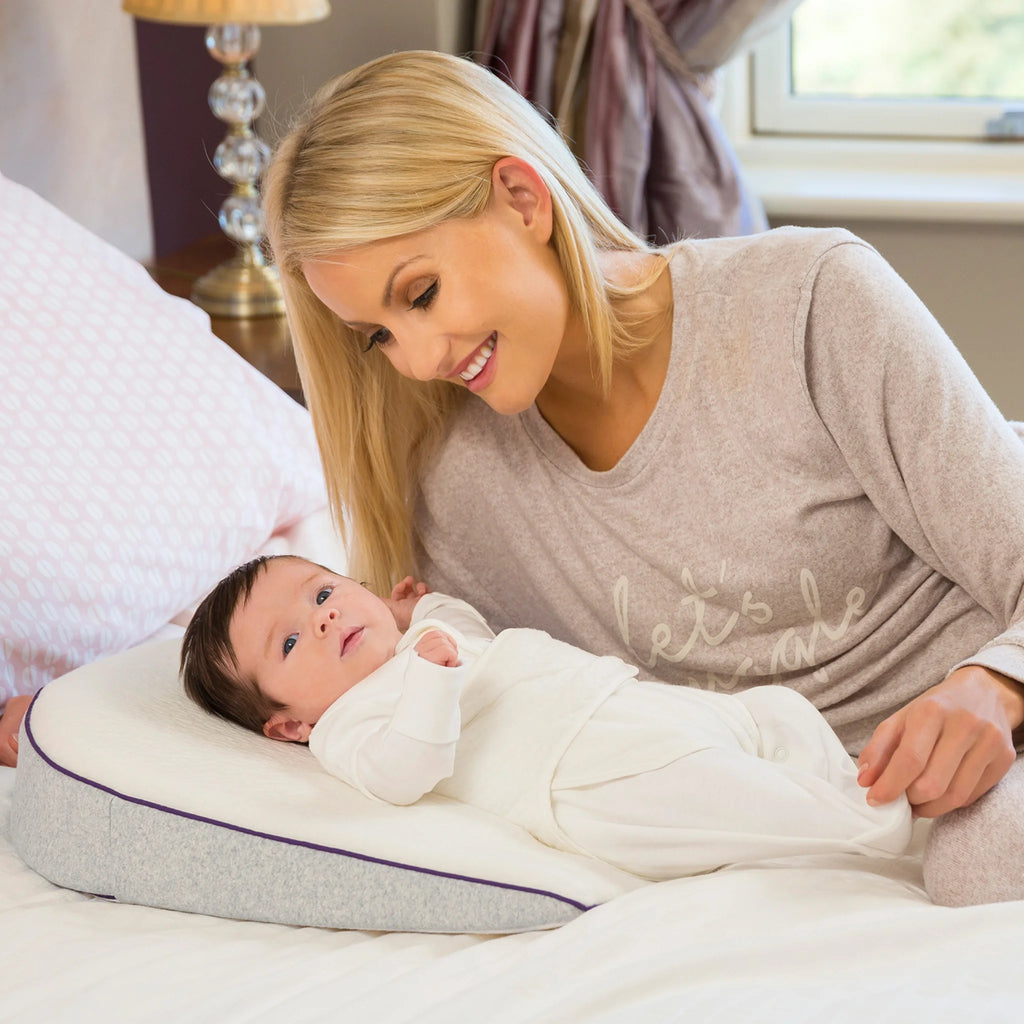 Smiling mother bonding with her baby lying comfortably on a Clevamama ClevaFoam Elevated Support wedge pillow, designed to reduce reflux, aid digestion, and provide safe elevated sleep support for newborns and infants.