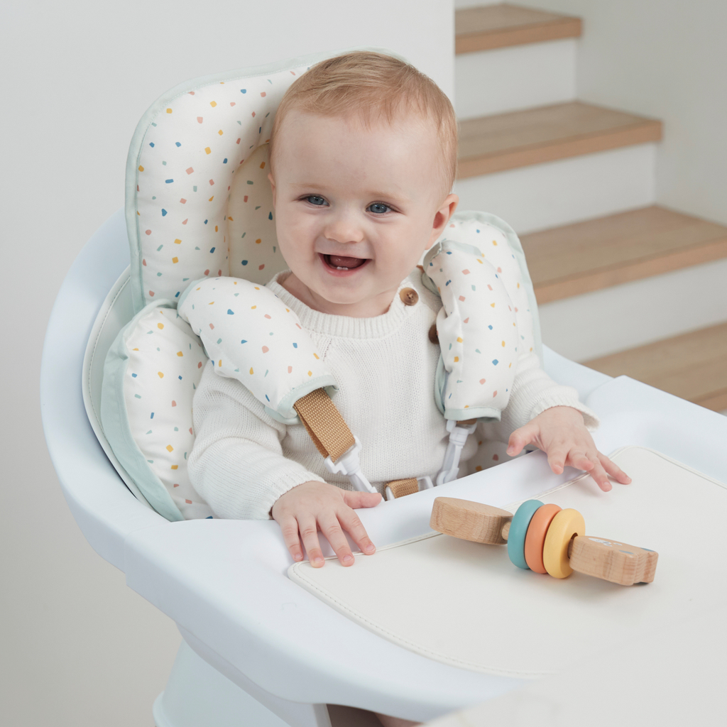 Smiling baby sitting in a high chair with padded support cushion and safety harness, soft cotton insert with colorful confetti print for comfort and protection.