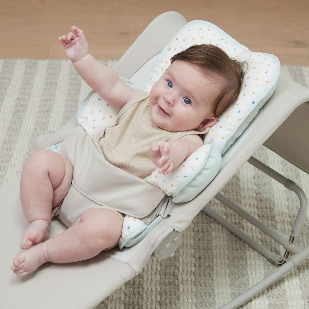 Baby sitting in a white baby bouncer with terrazzo print head and neck support on a wooden floor.