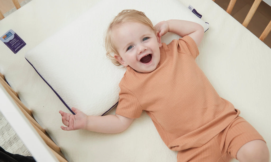 Toddler smiling lying on ClevaFoam Toddler Pillow and ClevaFoam Support Mattress in cot.