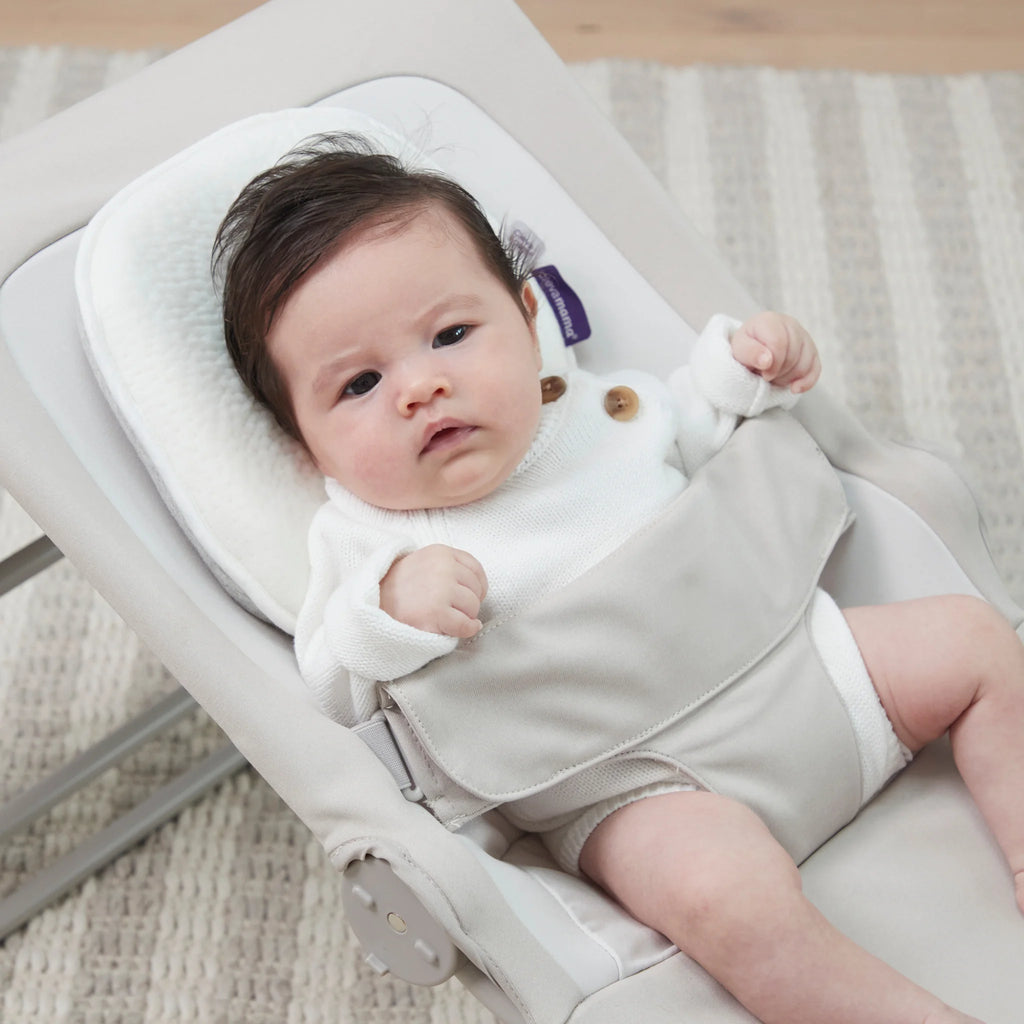 Baby in a white outfit lying on a beige baby bouncer with ClevaFoam Infant Pillow