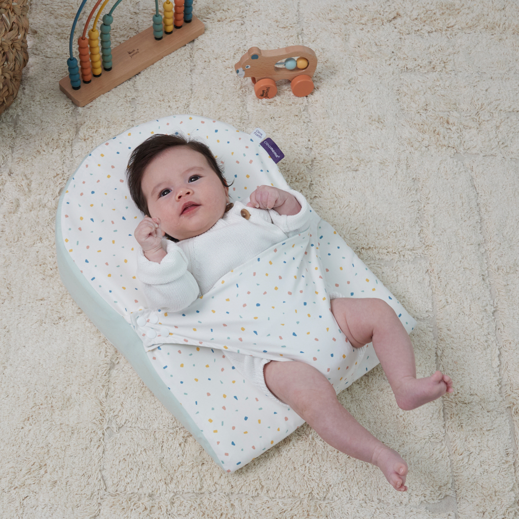 Baby lying on infant wedge pillow with adjustable swaddle wrap, breathable cotton newborn sleep positioner in colorful confetti print for comfort and safe support.