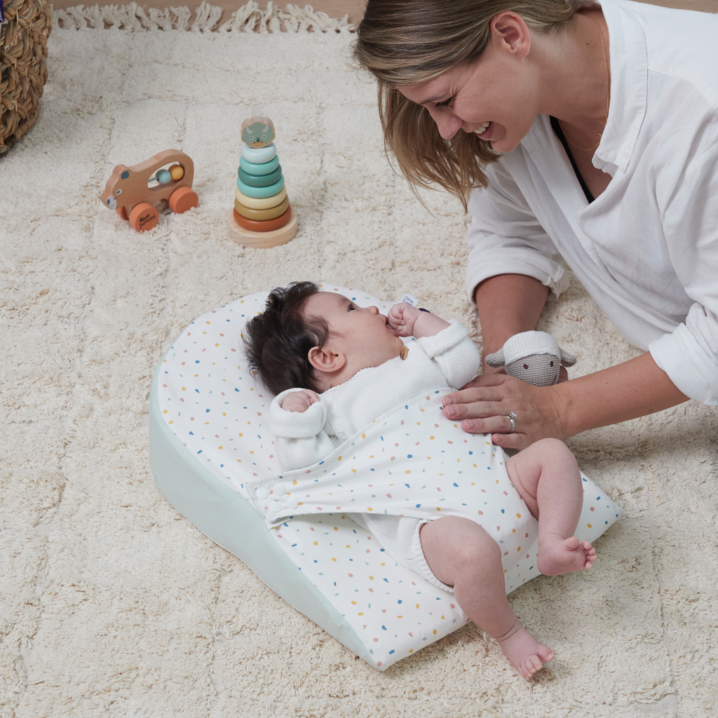Mother bonding with baby lying on newborn wedge pillow with swaddle wrap, breathable cotton infant sleep positioner in colorful confetti print for comfort and support.
