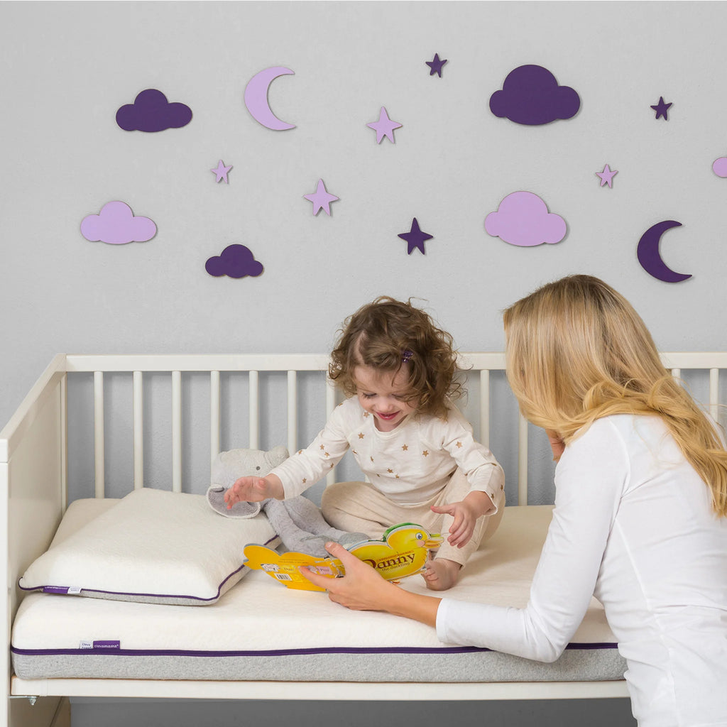 Mother reading bedtime story to toddler sitting on breathable hypoallergenic cot mattress in white crib, premium foam baby mattress for comfort, support and safe sleep.