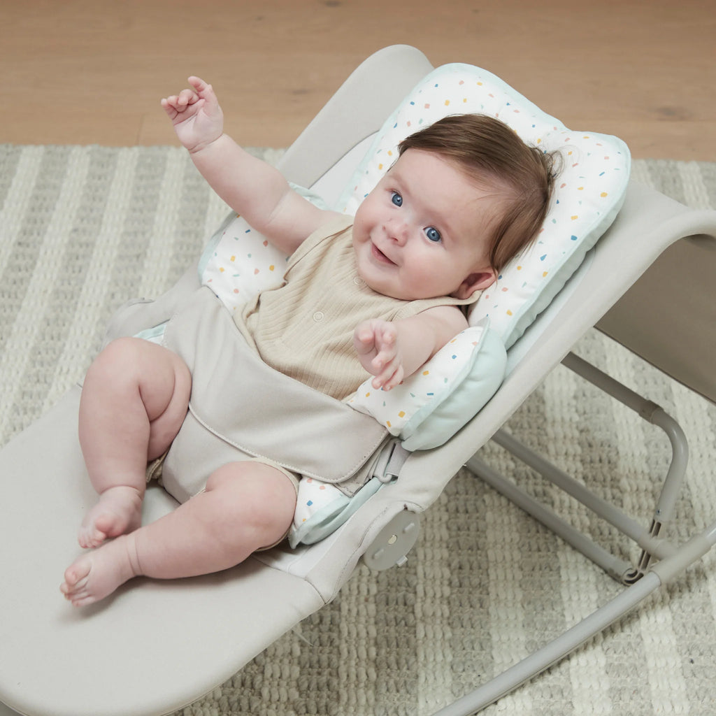 Baby in a white baby bouncer with a patterned head & neck support on a wooden floor.