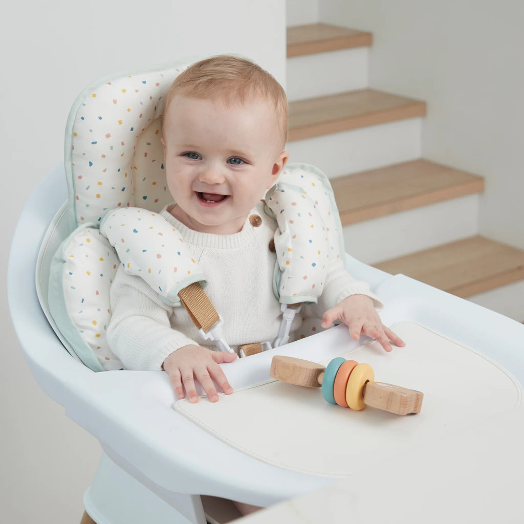 Baby sitting in a high chair with a toy, smiling, in a home setting with head and neck support.