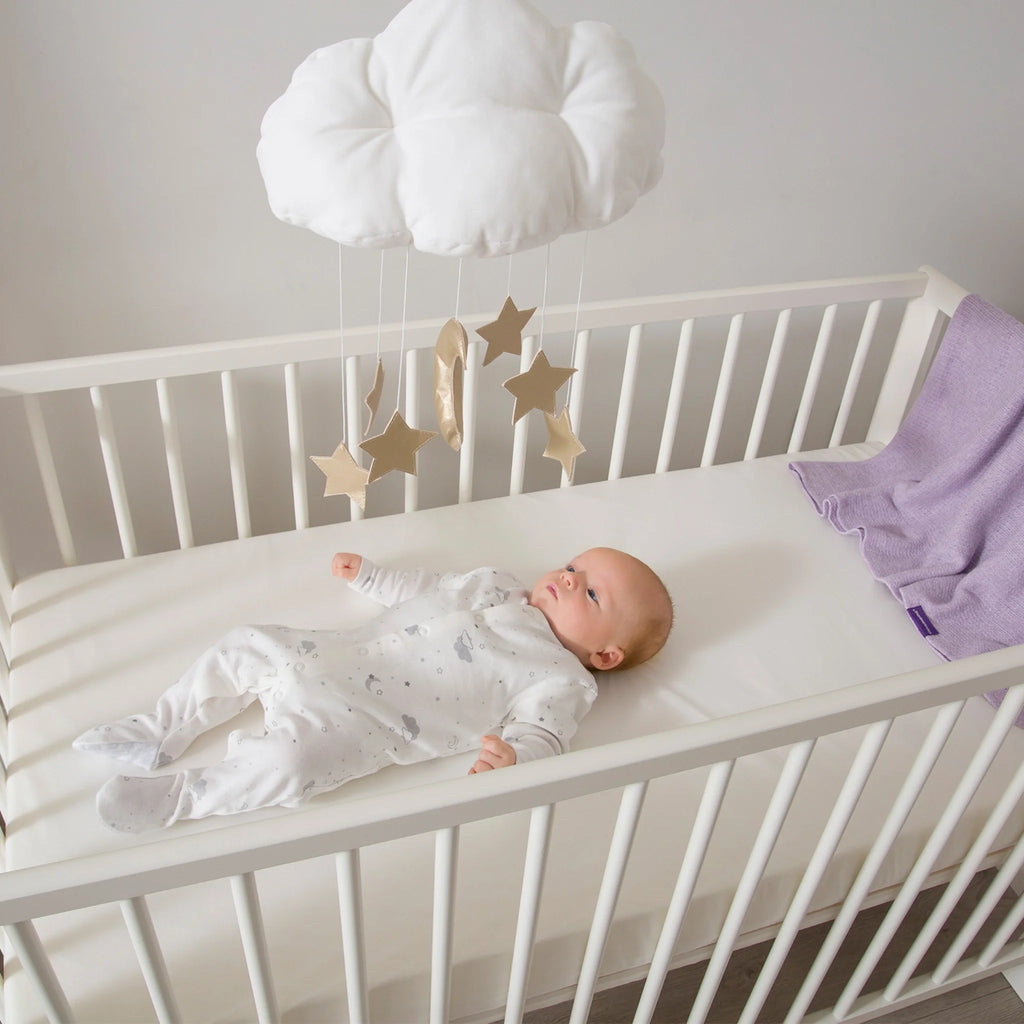 Baby lying on Clevamama waterproof breathable hypoallergenic cot mattress in white crib – premium foam crib mattress for infant comfort, support and safe sleep.