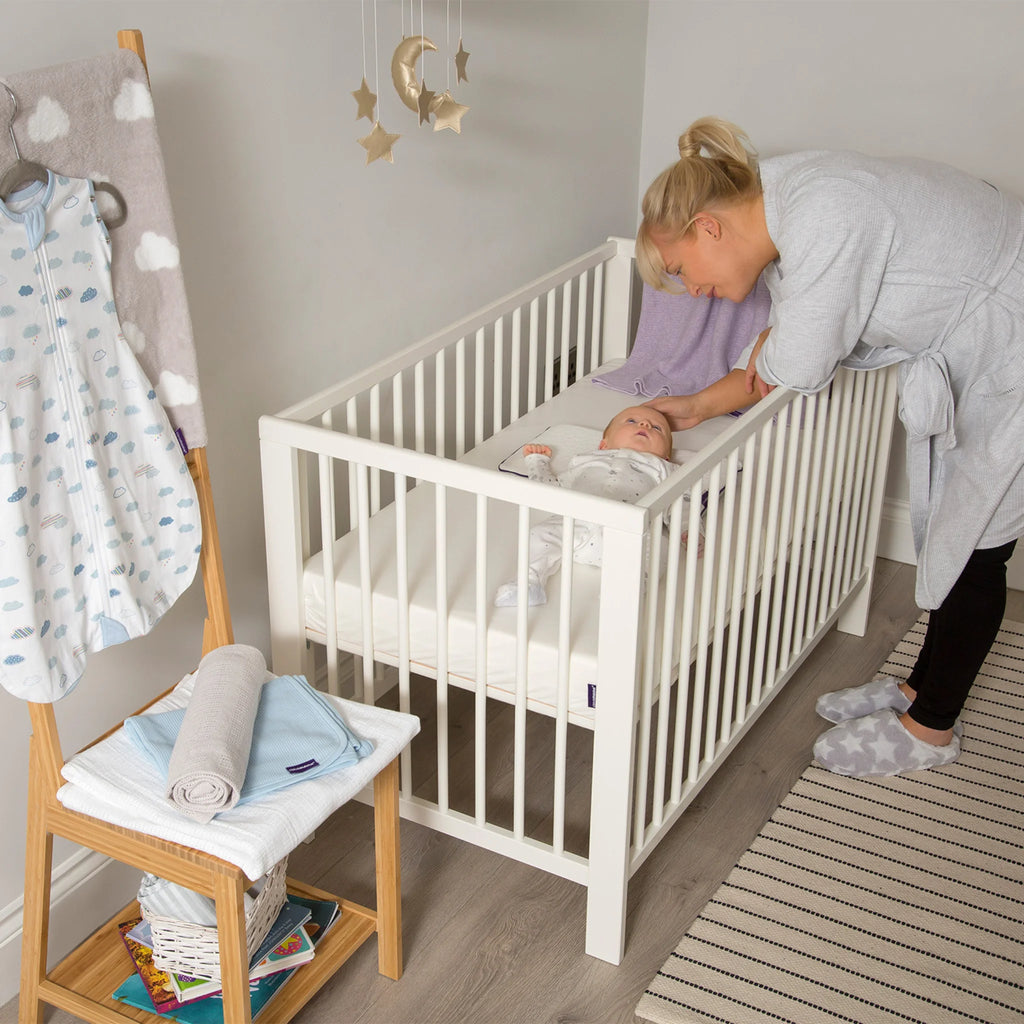 Mother soothing baby resting on Clevamama waterproof breathable hypoallergenic cot mattress in white crib – premium foam crib mattress for infant comfort, support and safe sleep.