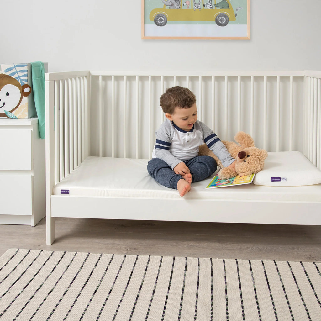 Toddler sitting in white cot bed reading with teddy bear on Clevamama waterproof breathable hypoallergenic mattress, premium foam crib mattress for comfort, support and safe sleep.